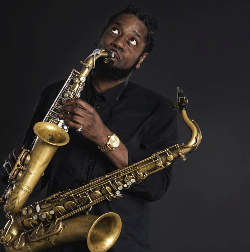 Soweto Kinch against a dark background holding multiple brass instruments